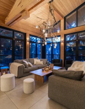 salon de la location chalet premium. Toit cathédrale, canapés, lustre spectaculaire et vue sur la foret depuis les grandes baies vitrées